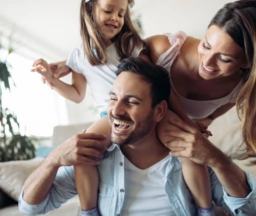 A man sits smiling with a young girl on his shoulders while a woman stands beside them, all appearing happy and playful indoors.