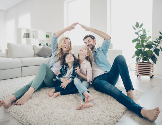 A family of four sits on a carpeted floor in a living room, smiling, with the parents forming a roof shape over their two children using their arms.