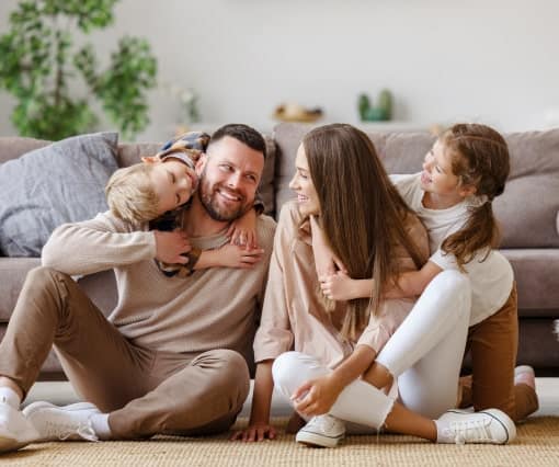 A family of four sits together on the floor in a living room, smiling and interacting playfully near a beige sofa.
