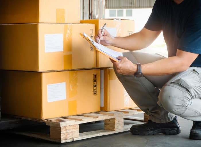 A person crouches next to a pallet stacked with cardboard boxes in a storage area, checking and recording packing information on a clipboard.