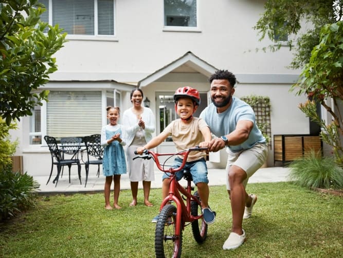 A man helps a young boy ride a bicycle on grass while two women watch and smile in front of a house.