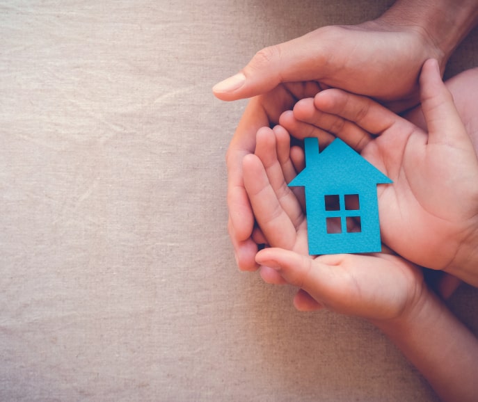 Two pairs of hands, one adult and one child, hold a blue paper cutout of a house together on a neutral fabric background.