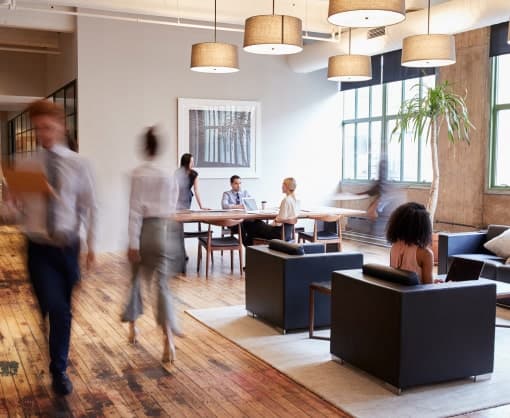 Modern open office space with people working and walking, featuring wooden floors, large windows, and contemporary furniture.