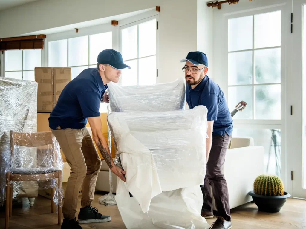 Residential and Commercial Moves: Two movers wearing blue uniforms and caps, carefully carry a large, plastic-wrapped chair into a bright living room filled with boxes and wrapped furniture.