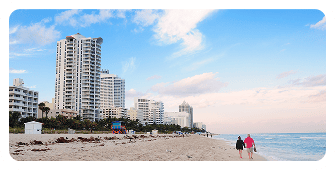 Two people walk along a sandy beach near the ocean, with tall modern buildings and palm trees lining the shore under a partly cloudy sky—capturing the relaxed vibe perfect for a Single Location Page.
