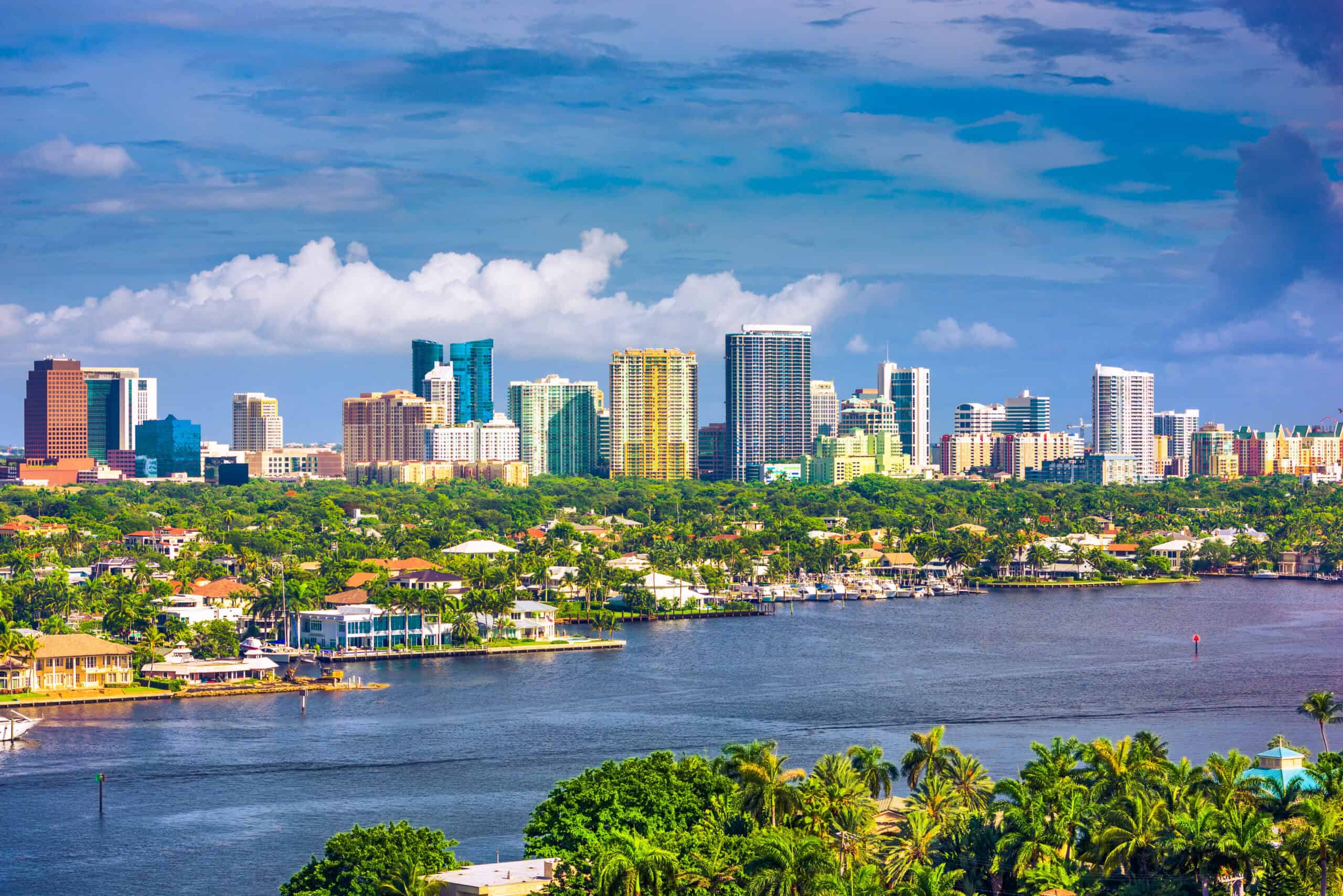 A view of a city skyline with high-rise buildings behind a wide river, palm trees, and residential neighborhoods highlights one of the most scenic urban locations under a partly cloudy sky.