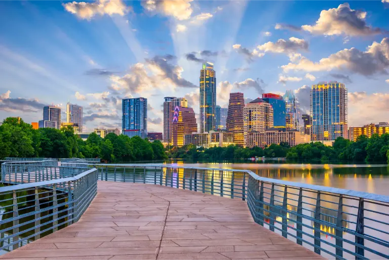 A view of the Austin, Texas skyline with tall buildings, trees, and a boardwalk over water under a partly cloudy sky with sun rays—perfect for those seeking trusted movers Austin relies on.