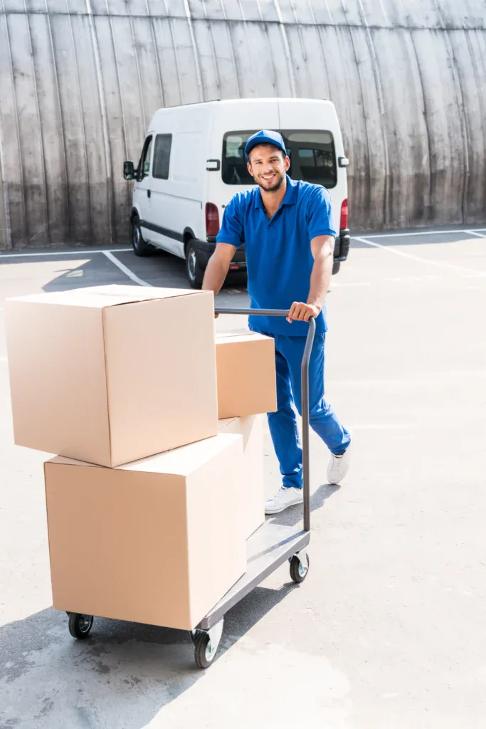A delivery worker representing one of White Room Moving Group's national moving companies pushes a cart with three large cardboard boxes in a parking lot.